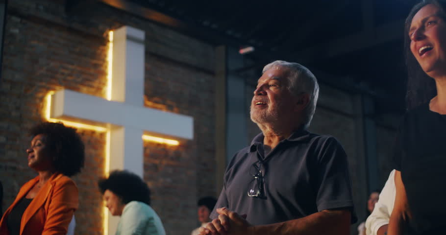 Elderly man standing in church with hands clasped, illuminated cross in background, diverse congregation worshipping in unity, expression of faith and spirituality