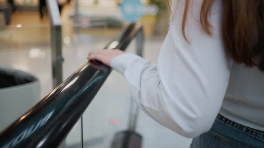 Close-up of lady with black polished nails tapping escalator handrail while descending, she wears a white blouse with long sleeves, blurred background showcases modern shopping mall interior