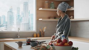 Shooting of young good-looking caucasian woman with towel on head preparing healthy breakfast in modern kitchen. Beautiful housewife in bathrobe cutting vegetables and looking at skyscrapers outside - Powered by Shutterstock - Get 15% off with code: PIKWIZARD15