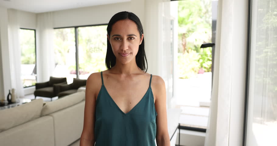 Happy beautiful young New Zealander woman posing at stylish home for head shot portrait, standing in living room, crossing arms at chest, getting cheerful, laughing, looking at camera