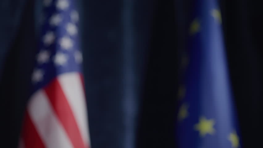 Close-up of handshake between male and female politicians in formal suits during international press conference, with American and European Union flags in background