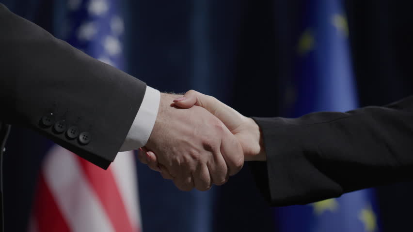 Close-up of handshake between male and female politicians in formal suits during international press conference, with American and European Union flags in background