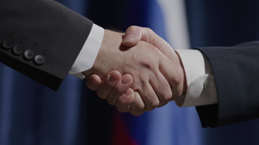 Close-up view of two male diplomats in formal suits shaking hands in front of Russian flag during political press conference