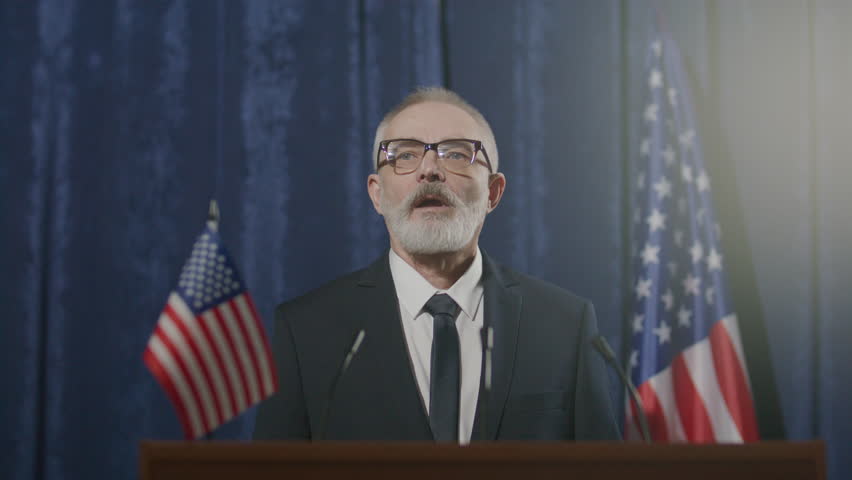 Senior male politician in formal suit and glasses delivering speech to press, standing at podium with American flag in background during official election campaign event
