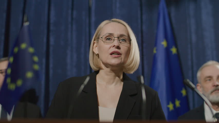Middle-aged female politician wearing formal attire and glasses, standing by podium with microphones and speaking to audience at European Union public event with flags and officials in background