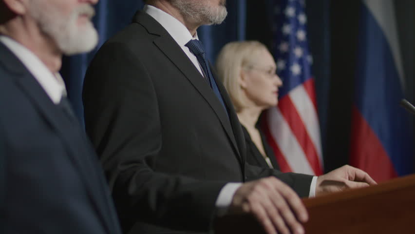 Middle-aged political leader standing with team on stage with American and Russian flags in background, making public announcement during press conference. Tilt-up shot