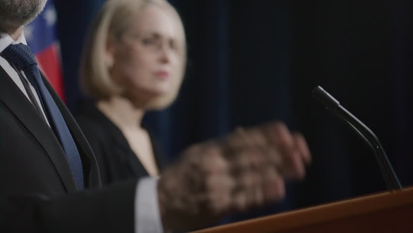Middle-aged male politician standing behind podium on stage with US flag in background, communicating with audience while participating in presidential debate