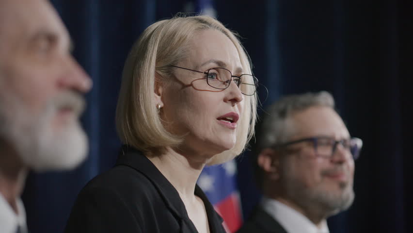 Female senator standing on stage with two male colleagues, delivering speech to audience during political press conference with US flag in background