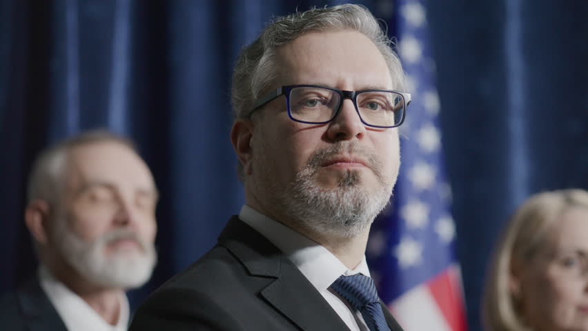 Middle-aged politician in suit and glasses looking confidently into camera during press conference with team members standing in front of American flag in background. Video portrait