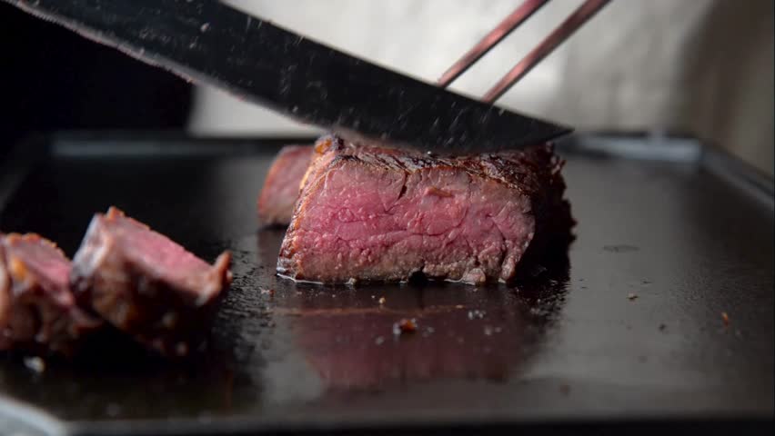 Close up of a chef cutting fatty beef with a knife while grilling a piece of meat on a grill pan in a restaurant kitchen. food close up zoom in