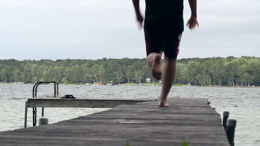 Teenage boy runs and jumps off the end of a wooden dock at the side of a lake.