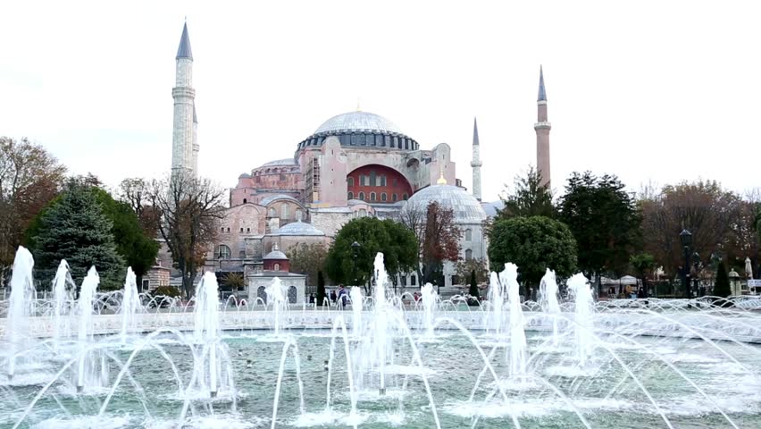 Fountain dances gracefully in foreground while Hagia Sophia stands tall in background. Clear blue sky enhances stunning architecture of this historic site. Visitors enjoy serene atmosphere.