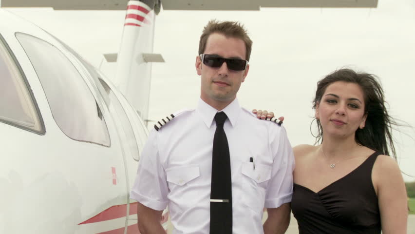 Pilot in uniform and young woman in a black dress pose next to an executive jet.