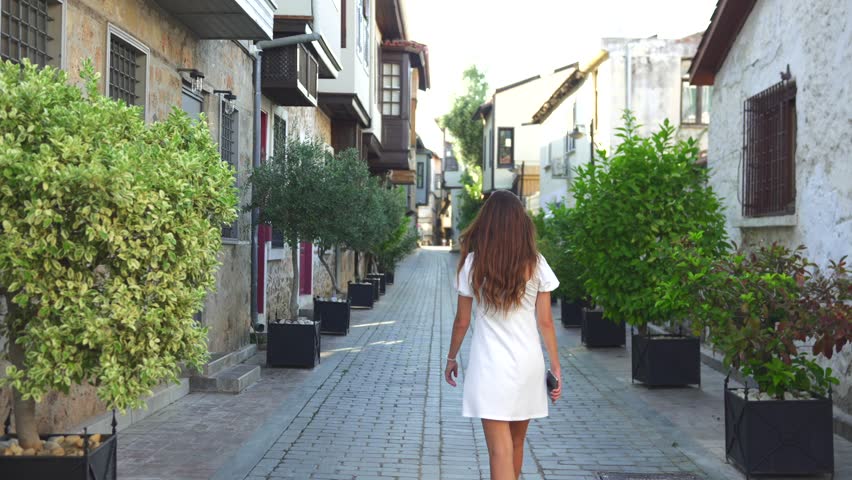 Young woman in white dress strolling through a cobblestone alley surrounded by greenery and historic stone houses in a quaint village. Antalya city, Turkey 2025 