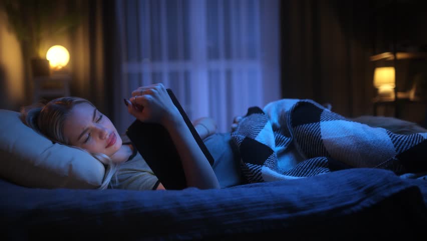 Tired young woman scrolling through social media on a tablet late at night while lying in bed, struggling to stay awake in a cozy dimly lit bedroom