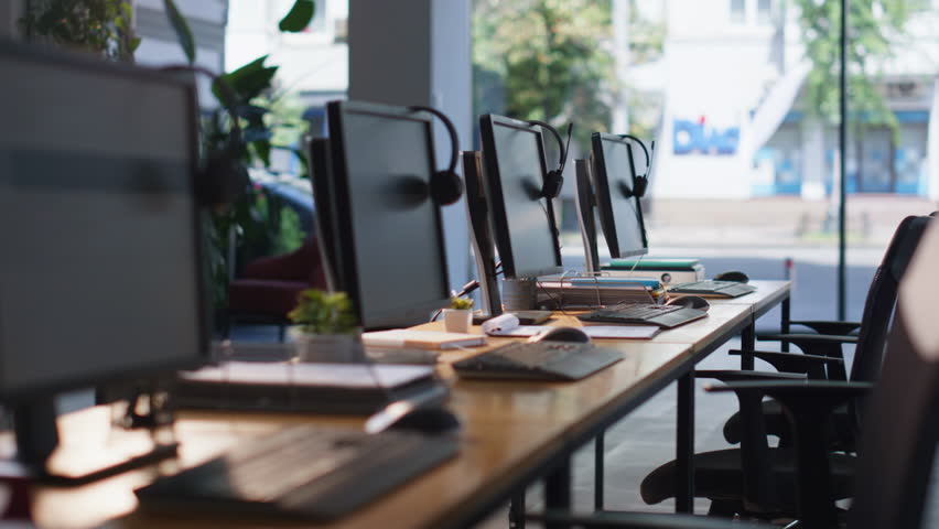 Empty office desks with computers and headphones at modern workspace. Organized call center workplace ready for teamwork and communication. Stylish corporate atmosphere for customer support providing.