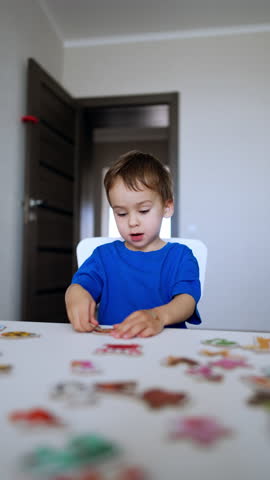 Smart cute toddler kid looks at the puzzles scattered on the desk in front of him. Kid picks the pieces playing a game. Vertical video.