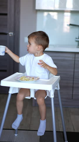 Toddler kid sitting at high chair drinks from a cup. Baby boy finishes drinking and gives a cup to a parent. Vertical video.