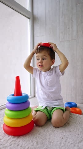 Happy Caucasian toddler sitting on the floor near the window. Cheerful kid plays with pyramid and puts the detail on his head. Vertical video.