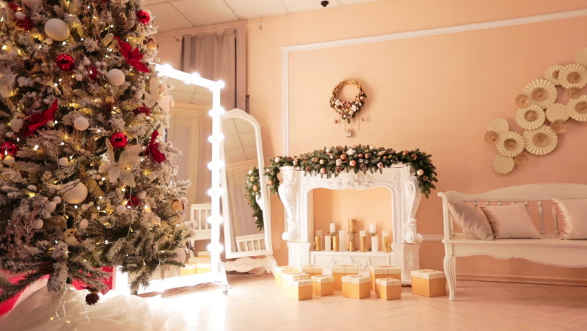 Coming closer to an artificial mantelpiece decorated with candles, garlands of fir trees, gift boxes and wreath above. Christmas photo zone in modern studio.