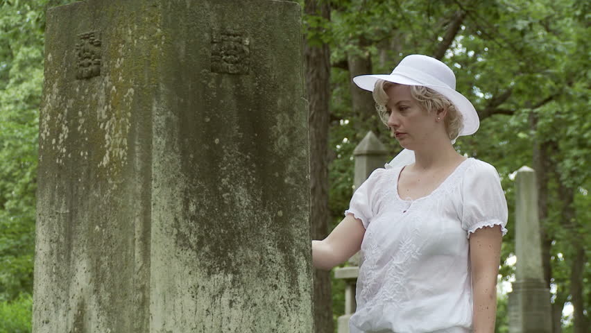 Beautiful, sad woman dressed in white visits a headstone in a graveyard. Mid
