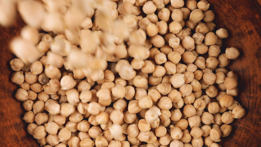 Chickpea seeds are poured into a brown ceramic bowl on a wooden oak board background. Top view. Slow motion.