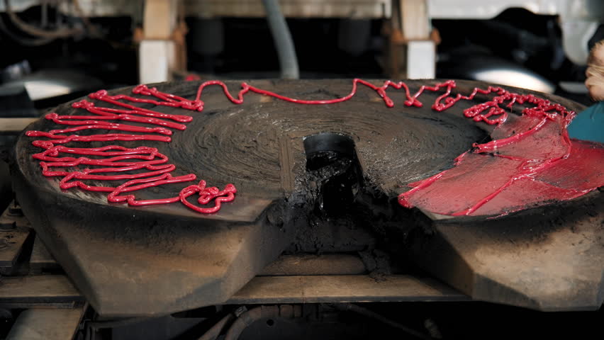 Mechanic covering the fifth wheel coupling on a truck with grease for lubrication, with red grease or marking compound applied around the edges. Truck is in repair shop during the inspection.