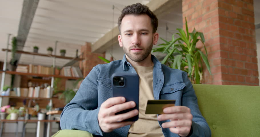 Male on Sofa looking at Smart phone and holding Credit card for E-commerce Purchase