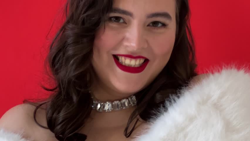 Horizontal video closeup. Young adult white brunette pin up woman with bare shoulder, scarlet lip, fluffy fur cape, diamond necklace smiles and looks at camera on red. Concept of 8 March, luxury