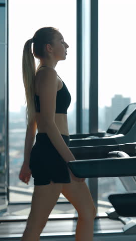 Young woman in shorts walks on treadmill in sports club against window. Confident female athlete prepares body for intensive workout with cardio activity in gym vertical video