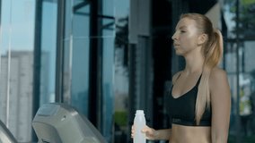 Portrait of woman with ponytail drinking water from bottle walking on treadmill in gym. Athletic young female in black sports bra stays hydrated during cardio workout looking at window - Powered by Shutterstock - Get 15% off with code: PIKWIZARD15