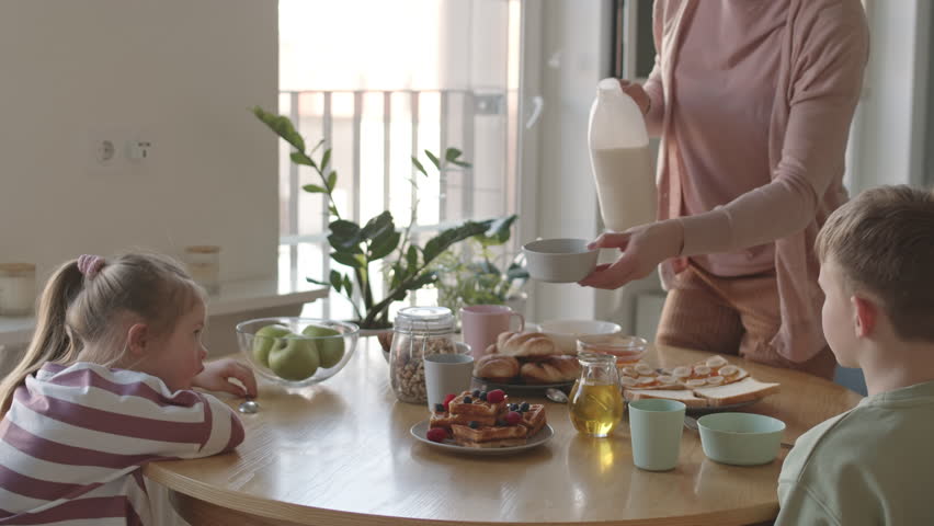 Lockdown of two kids sitting at dining table and waiting for their unrecognizable mommy standing at table and pouring fresh milk into bowls