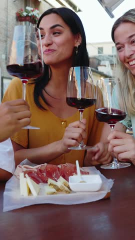 Vertical HD video of three young multiracial women toasting red wine sitting outside at restaurant table during summer holiday. Female friendship and vacation lifestyle concept.