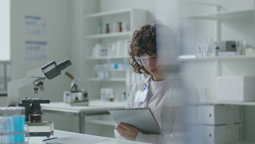 Young female scientist in white coat and safety glasses sitting at desk with microscope, using digital tablet during workday in modern medical laboratory - Powered by Shutterstock - Get 15% off with code: PIKWIZARD15