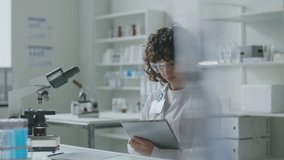 Young female scientist in white coat and safety glasses sitting at desk with microscope, using digital tablet during workday in modern medical laboratory - Powered by Shutterstock - Get 15% off with code: PIKWIZARD15