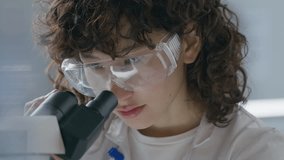 Young female scientist in safety goggles and white coat, adjusting focus knob and looking through microscope during laboratory research. Close-up, tilt-up shot - Powered by Shutterstock - Get 15% off with code: PIKWIZARD15