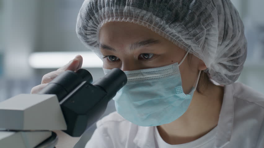 Asian female scientist wearing protective mask, gloves and cap observing sample through microscope, performing detailed research in medical laboratory. Close-up view