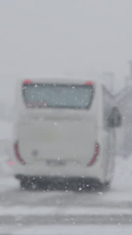 SLOW MOTION, DOF: White bus slows down at a bus stop during a snowy winter day. Bad weather gathers over the city streets and covers them in snow. Tourist coach stops due to bad weather conditions.