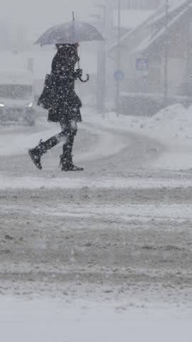 SLOW MOTION, DOF: Unrecognizable woman crosses the snowy street during a blizzard on a cold winter day. Unknown pedestrian walking across the empty snowy road in the middle of an intense snowstorm.