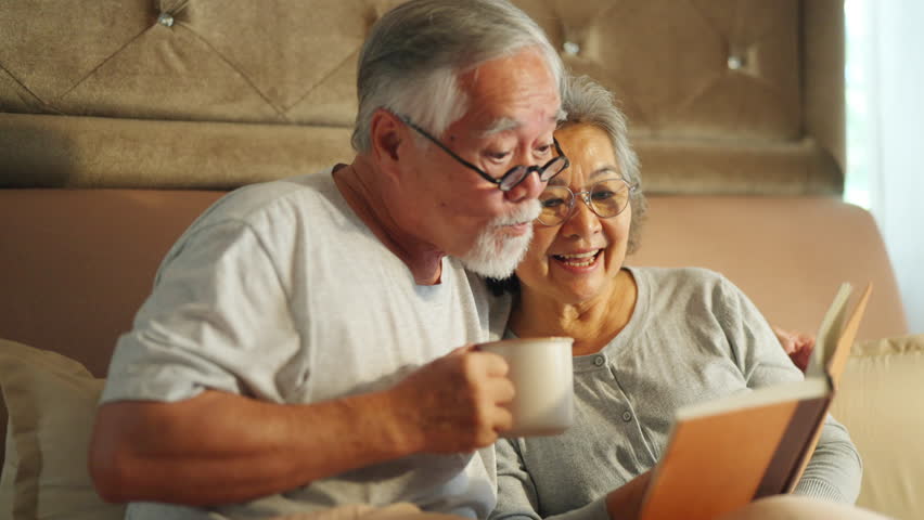 Happy Asian family senior couple relaxing and reading a book together on the bed in bedroom. Elderly husband and wife taking care each other. Retired people healthcare and mental health concept.
