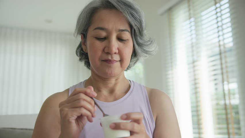 Asia adult people middle age happy old woman eat greek yogurt cup weight loss protein gut health care diet detox sit at home sofa elder aging asian aged female. Fresh medical food relief meal enjoy.