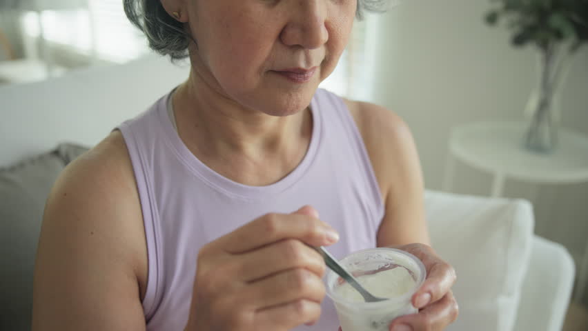 Asia adult people middle age happy old woman eat greek yogurt cup weight loss protein gut health care diet detox sit at home sofa elder aging asian aged female. Fresh medical food relief meal enjoy.