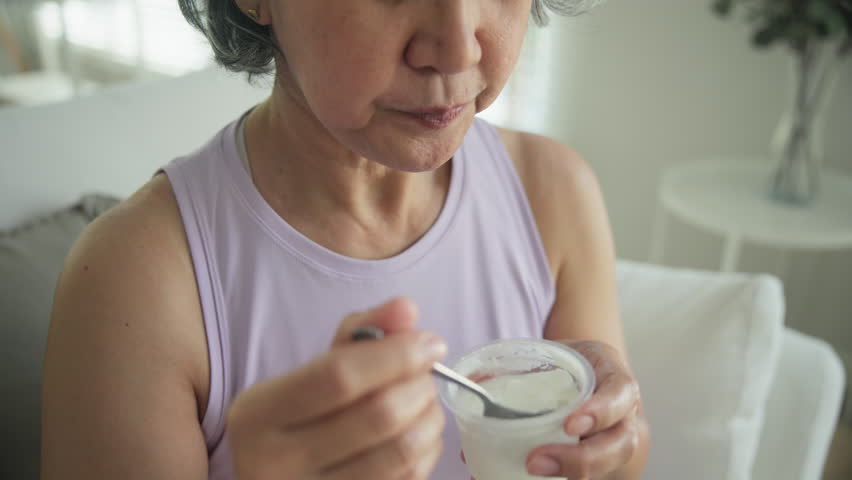 Asia adult people middle age happy old woman eat greek yogurt cup weight loss protein gut health care diet detox sit at home sofa elder aging asian aged female. Fresh medical food relief meal enjoy.