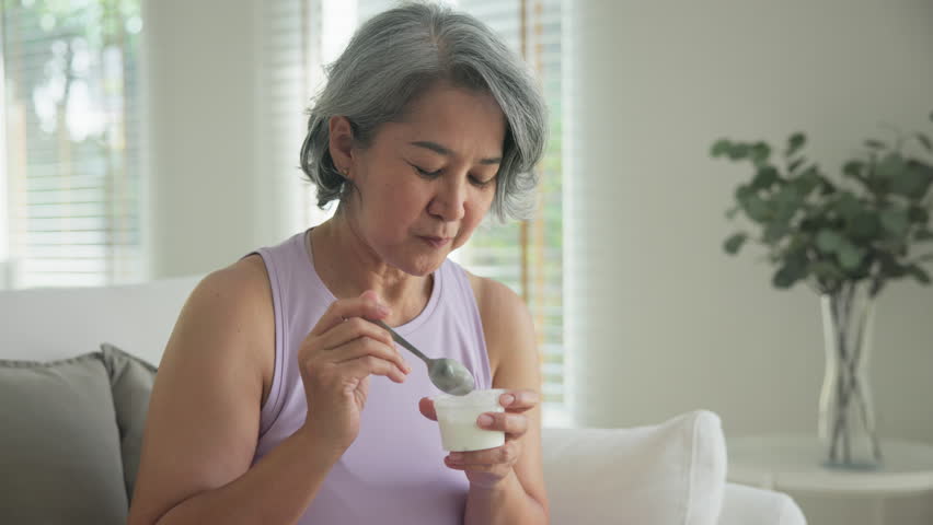 Asia adult people middle age happy old woman eat greek yogurt cup weight loss protein gut health care diet detox sit at home sofa elder aging asian aged female. Fresh medical food relief meal enjoy.