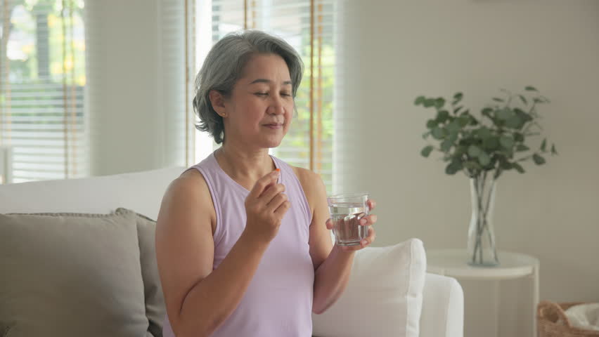 Asia adult aged people middle age happy old woman smile drink vitamin d c e k zinc omega 3 fish oil water pill reduce hot flash dry skin health care sit at home sofa elder aging asian. Take diet food.