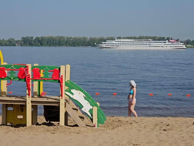 Woman is walking on the beach near a cruise boat. The boat is large and white. There is a playground in the background