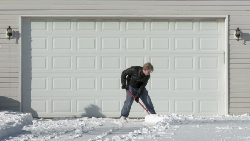 Young man digs away snow from in front of a garage in the Mid West, USA. Wide