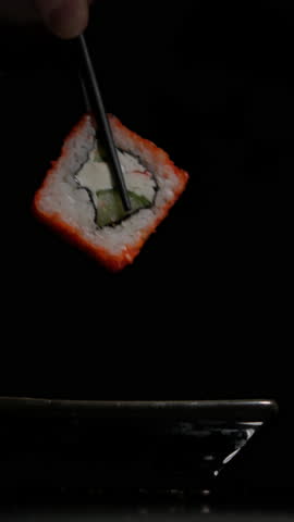 Sushi roll dipped in soy sauce on black background close-up vertical video. Man eats sushi roll with chopsticks while soaking it in sauce.