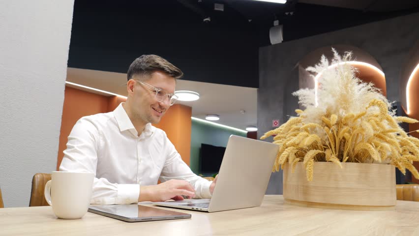Man in white shirt uses tablet while sitting at wooden table in contemporary workspace.
