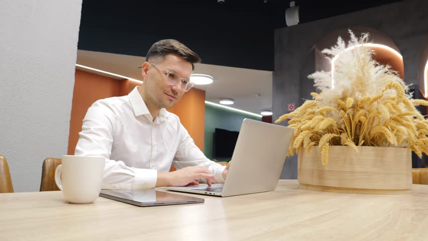 Man in white shirt uses tablet while sitting at wooden table in contemporary workspace.
